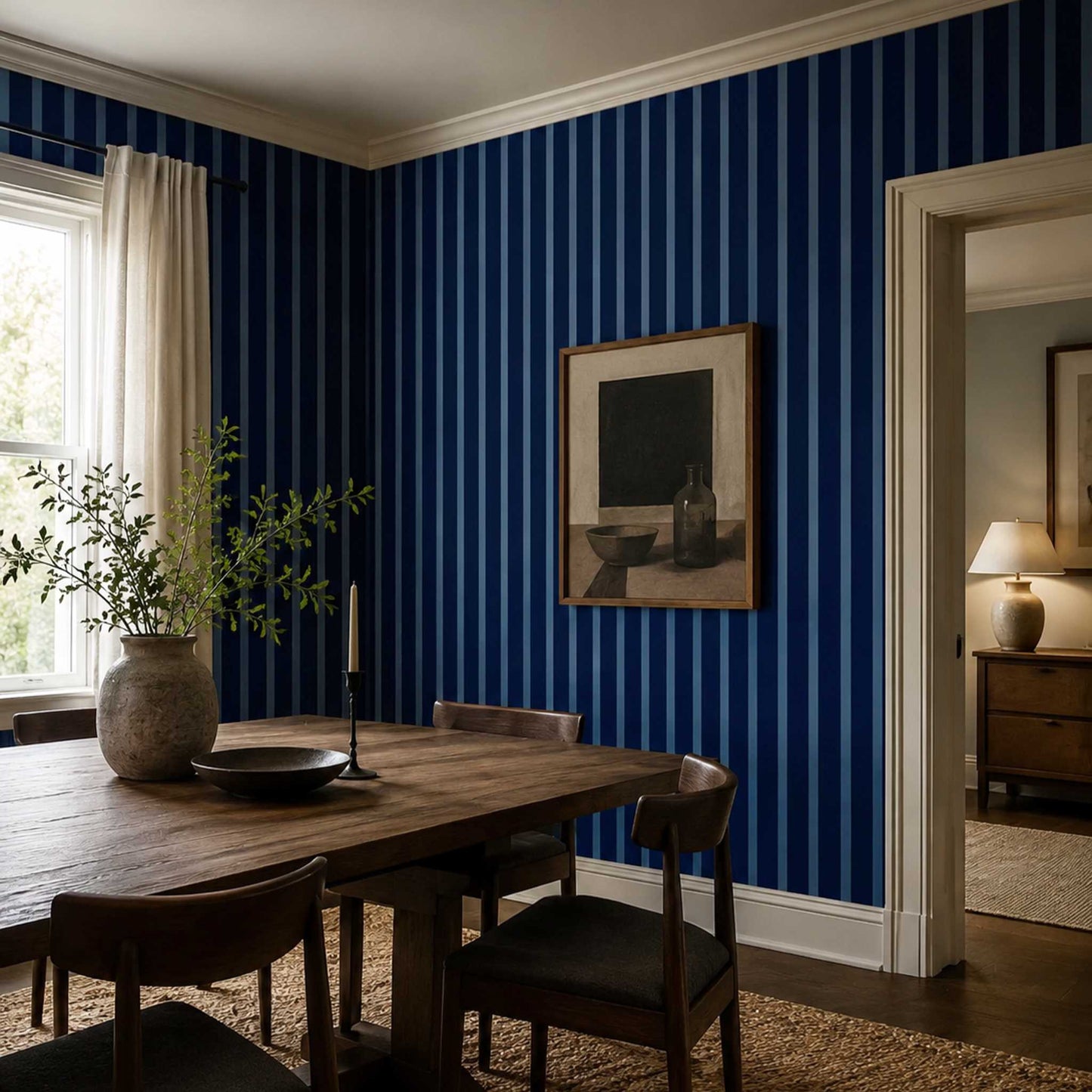 A dining room corner with bold but classic navy and sapphire blue vertical striped wallpaper, a wooden dining table, dark wood chairs, a large vase with greenery, and framed wall art.
