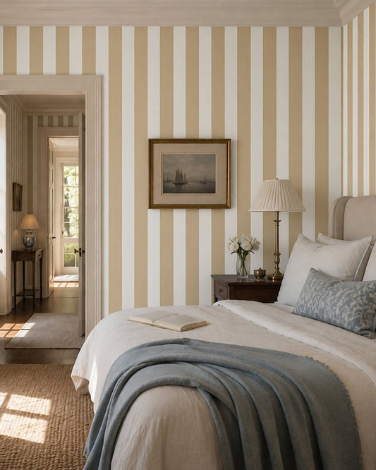 A traditionally styled bedroom featuring vertical tan and white striped wallpaper, a neatly made bed with white linens and a blue-gray throw blanket, a bedside lamp, and a framed antique sail boat print on the wall.